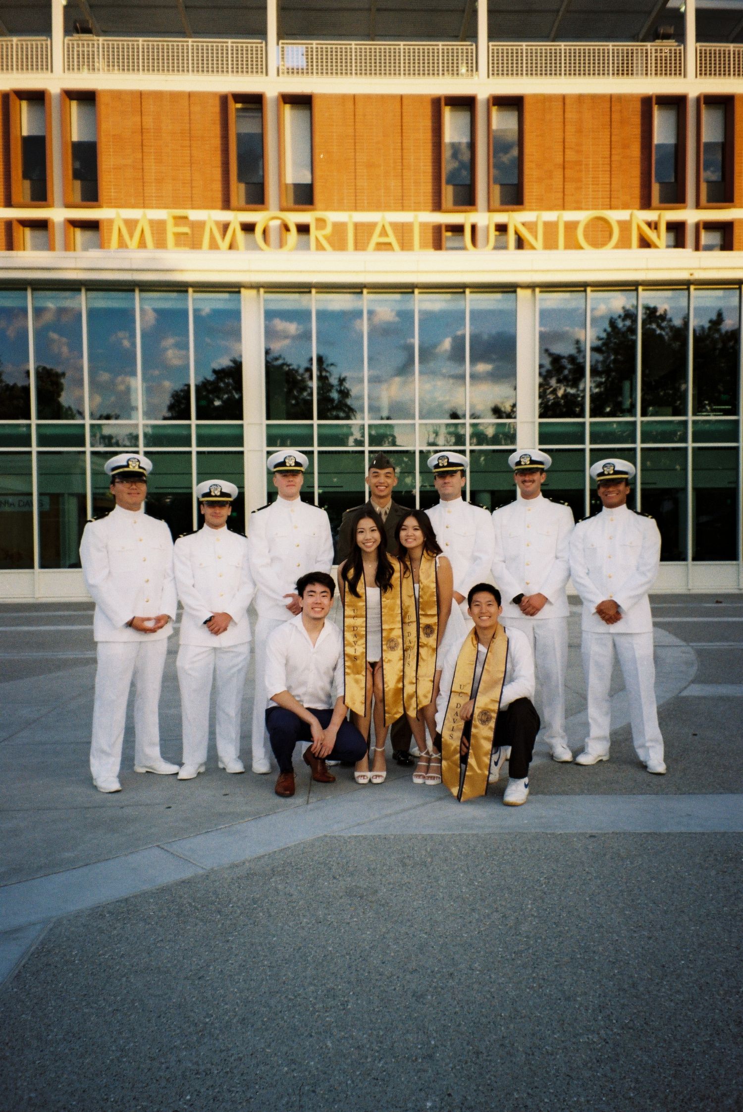A group photo in front of a Memorial Union building with graduates in white uniforms and gold stoles.