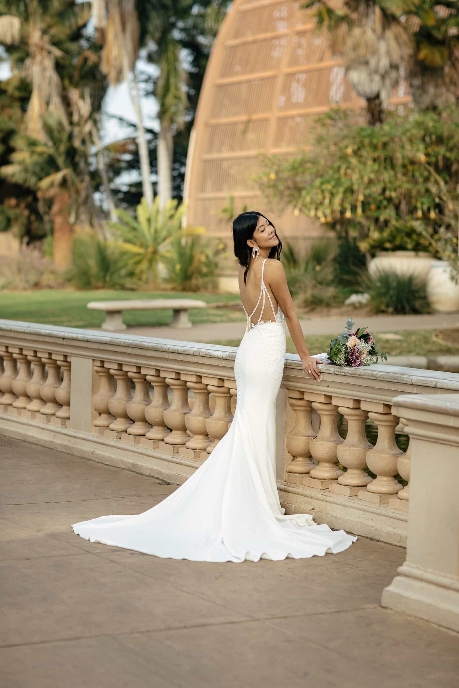 Bride in sleek white mermaid wedding gown with train poses at stone balustrade in lush garden setting.