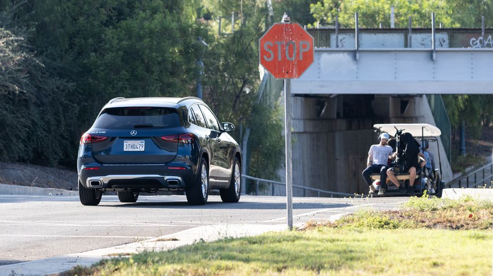 A dark blue car drives past a stop sign near an underpass while golf cart is visible in the background.