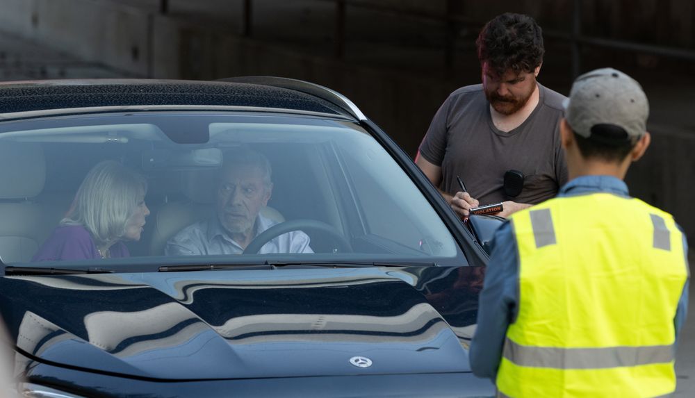 A worker in a high-visibility vest speaks with occupants of a dark vehicle.