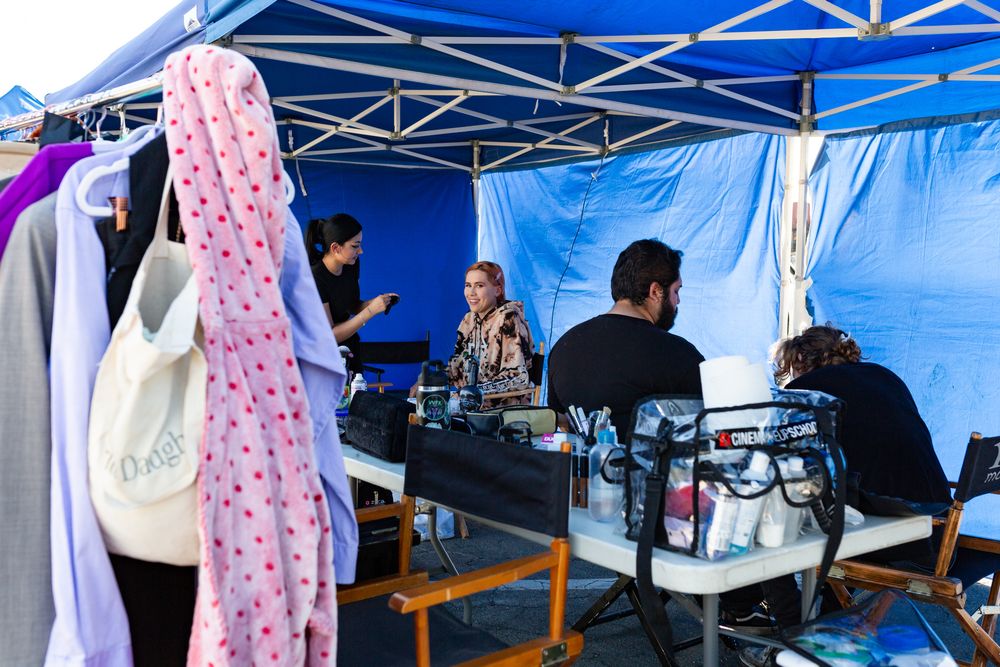 Production crew working under a blue tent with clothing racks and equipment visible.