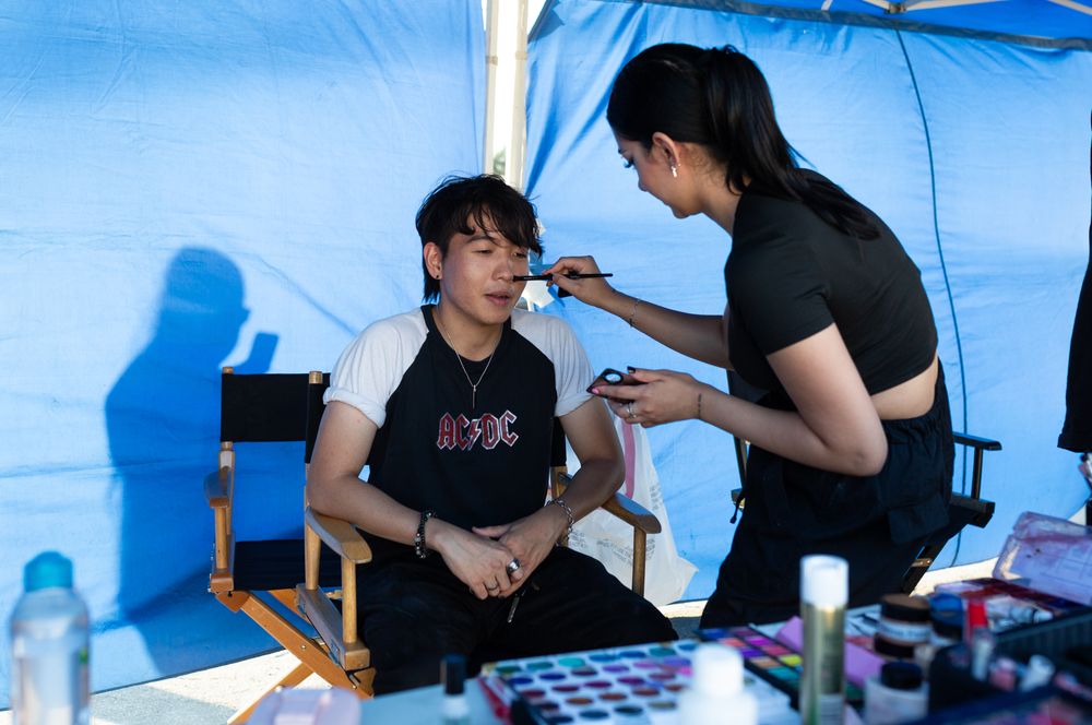 Two people in a makeup session under a blue tent during an outdoor production.