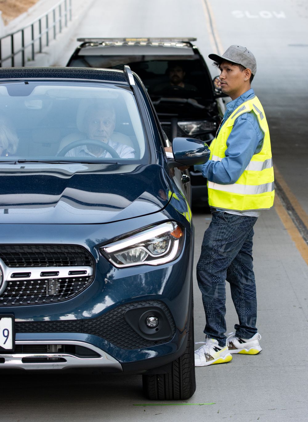 Worker in safety vest interacts with driver of blue Mercedes SUV in parking area.
