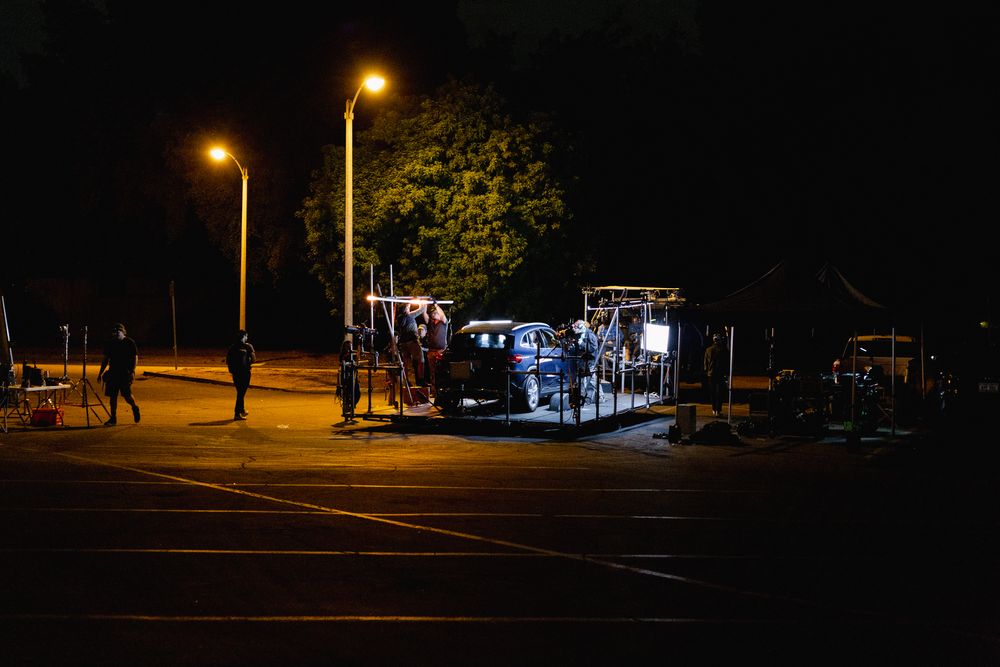 Empty parking lot at night illuminated by street lamps with buildings in background.