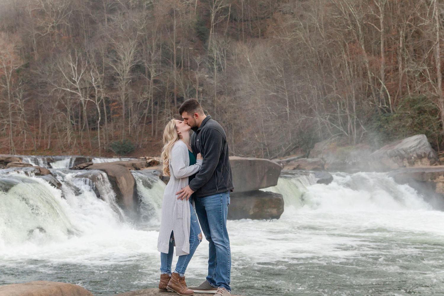 Couple shares intimate moment in front of rushing waterfall on cloudy day.