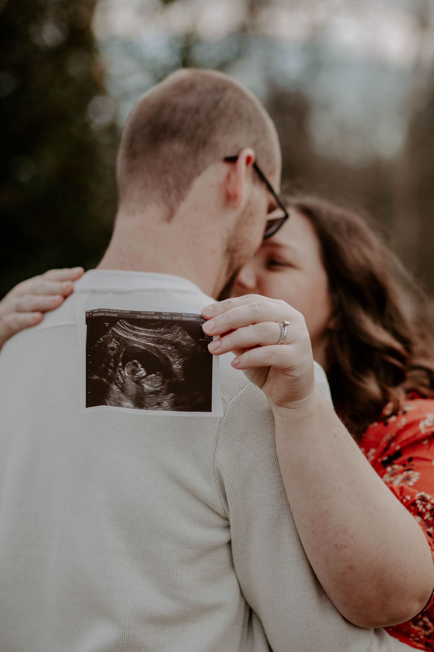 Maternity photos- A pregnant woman holds her ultrasound photo with her husband's arms wrapped around her.
