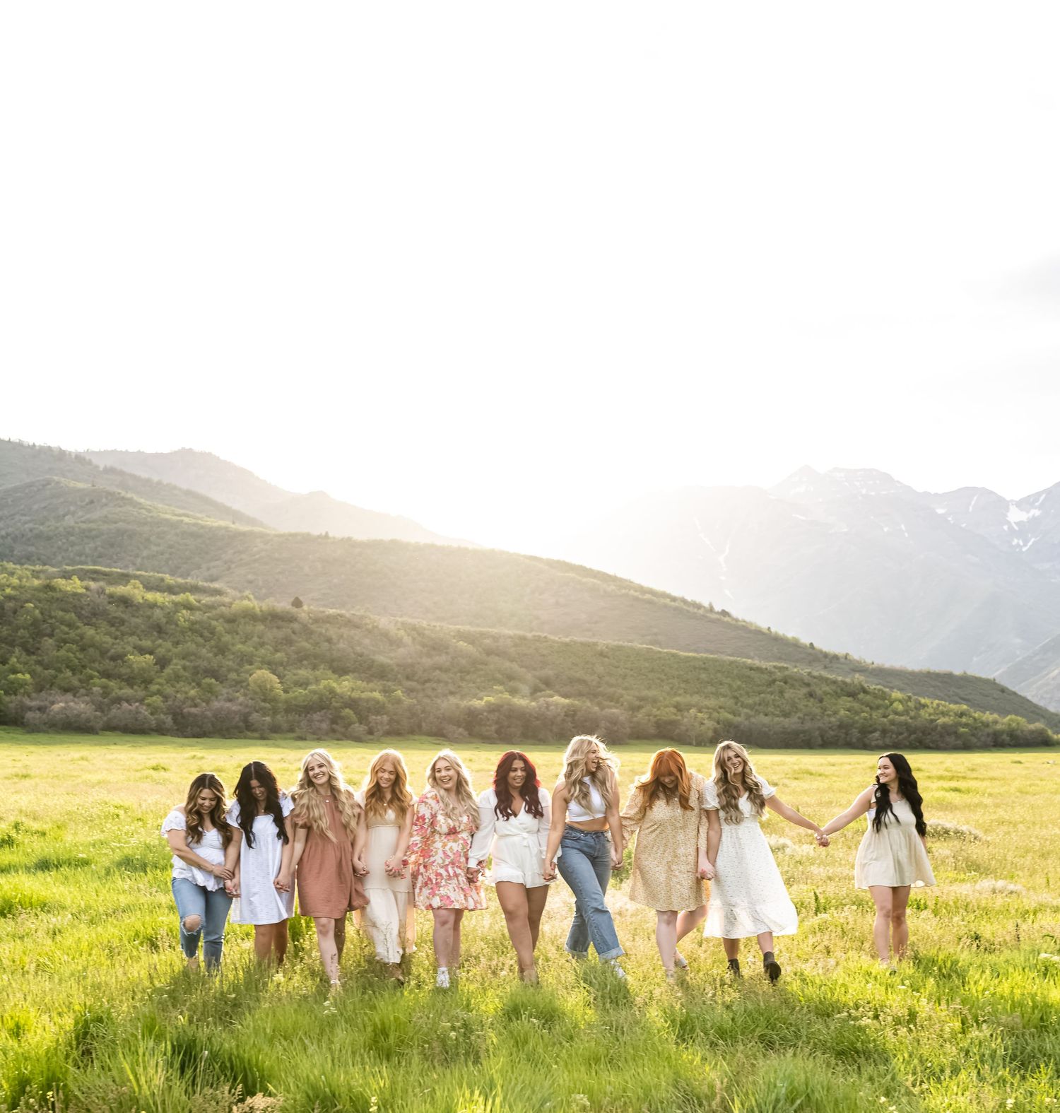 A group of women in coordinated outfits walking together through a sunlit meadow with mountains in the background.