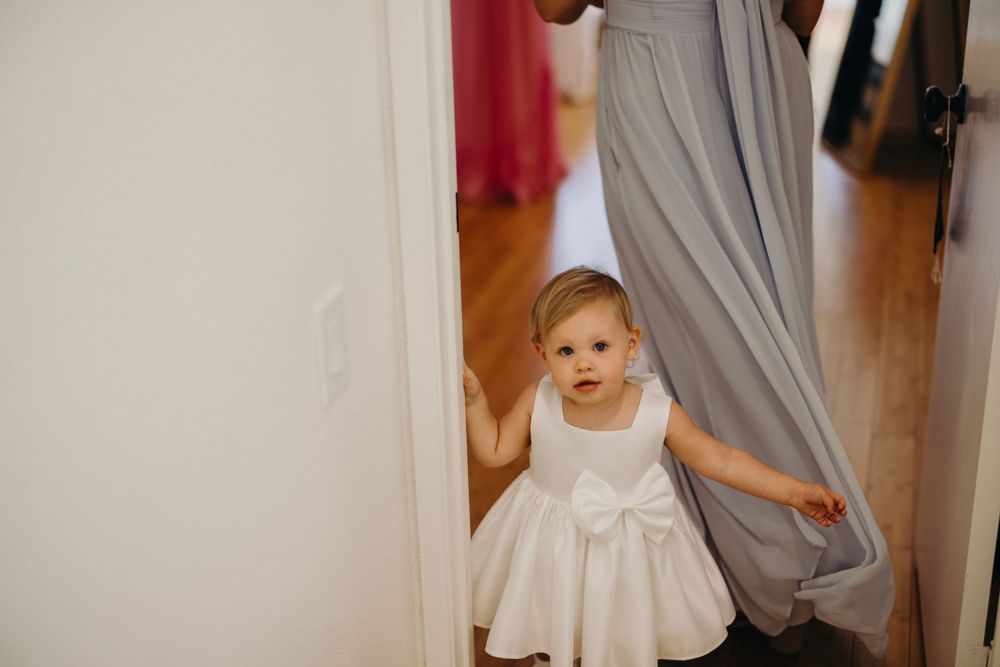 A small child in a white dress peeks around a doorway during wedding preparations.