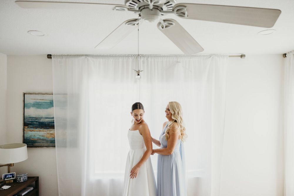 Two people in light-colored dresses stand by a window with white curtains and ceiling fan.
