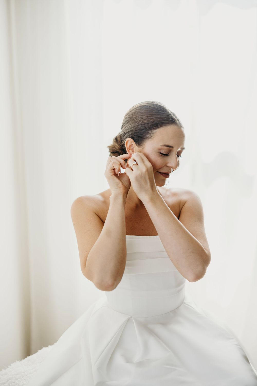 A bride in a classic white dress puts on earrings during wedding preparations.