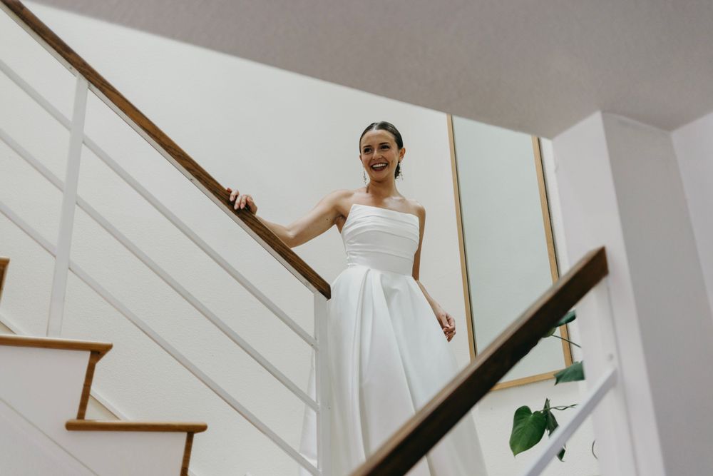 A person in a white wedding dress stands elegantly on a staircase with white walls and wooden railings.