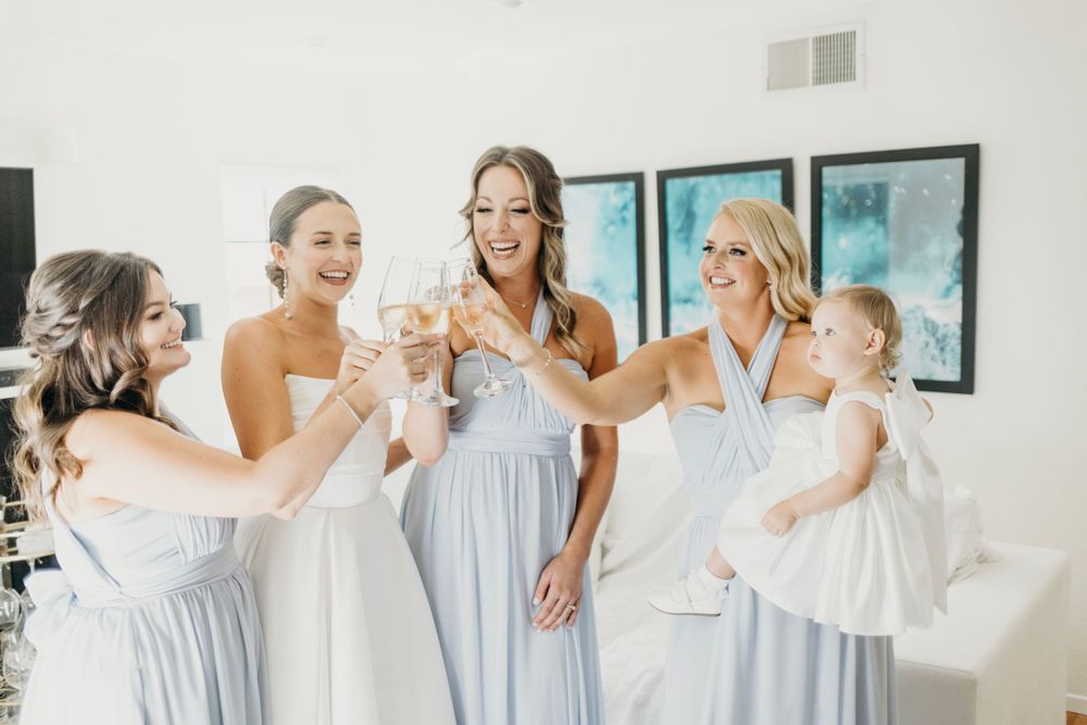 Bridesmaids in pale blue dresses share a celebratory toast in a bright indoor setting.