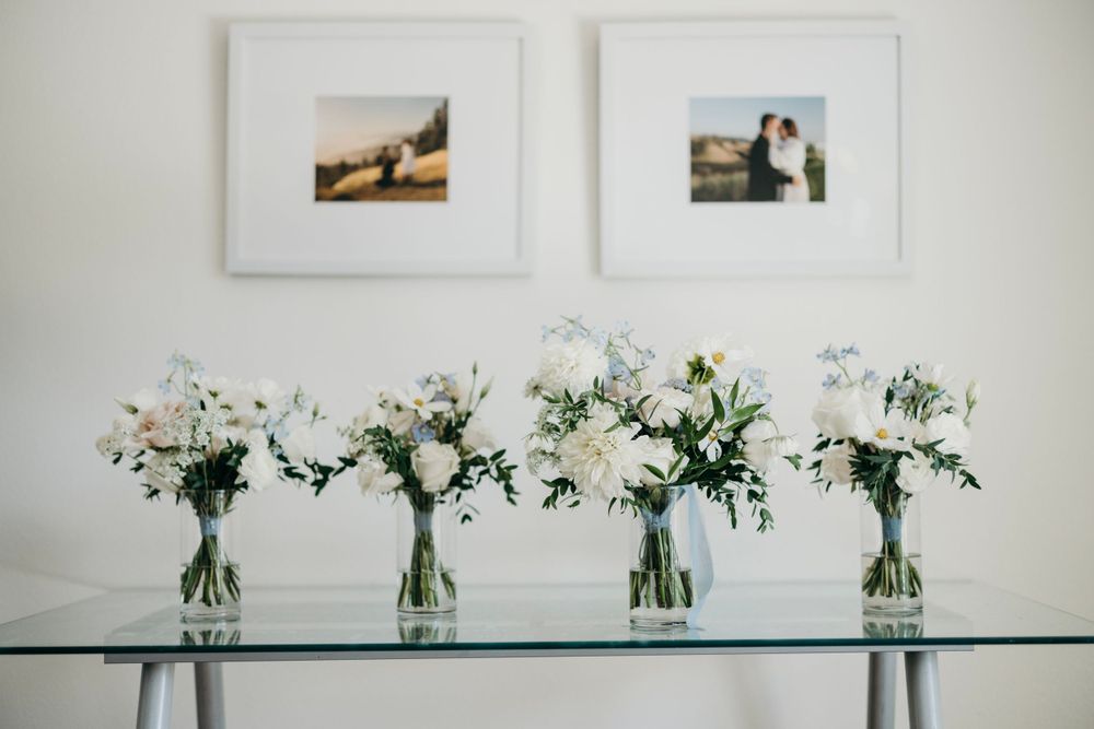 Four elegant floral arrangements with white blooms displayed in glass vases on a reflective table.