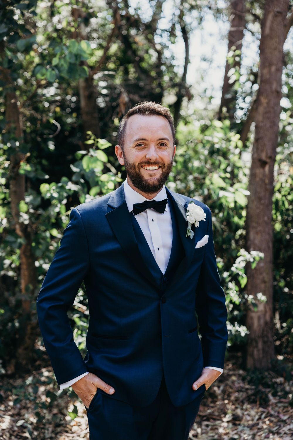 A groom in a navy tuxedo poses outdoors among trees and natural greenery.
