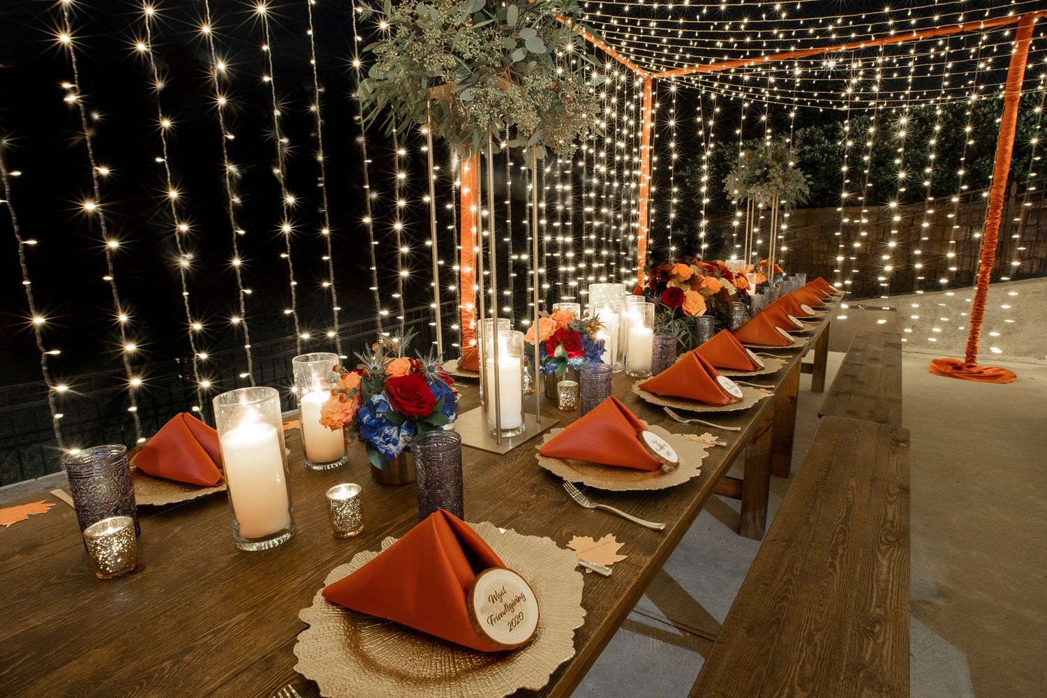 Elegant wedding reception table setting with string lights and orange napkins in a rustic outdoor setting.