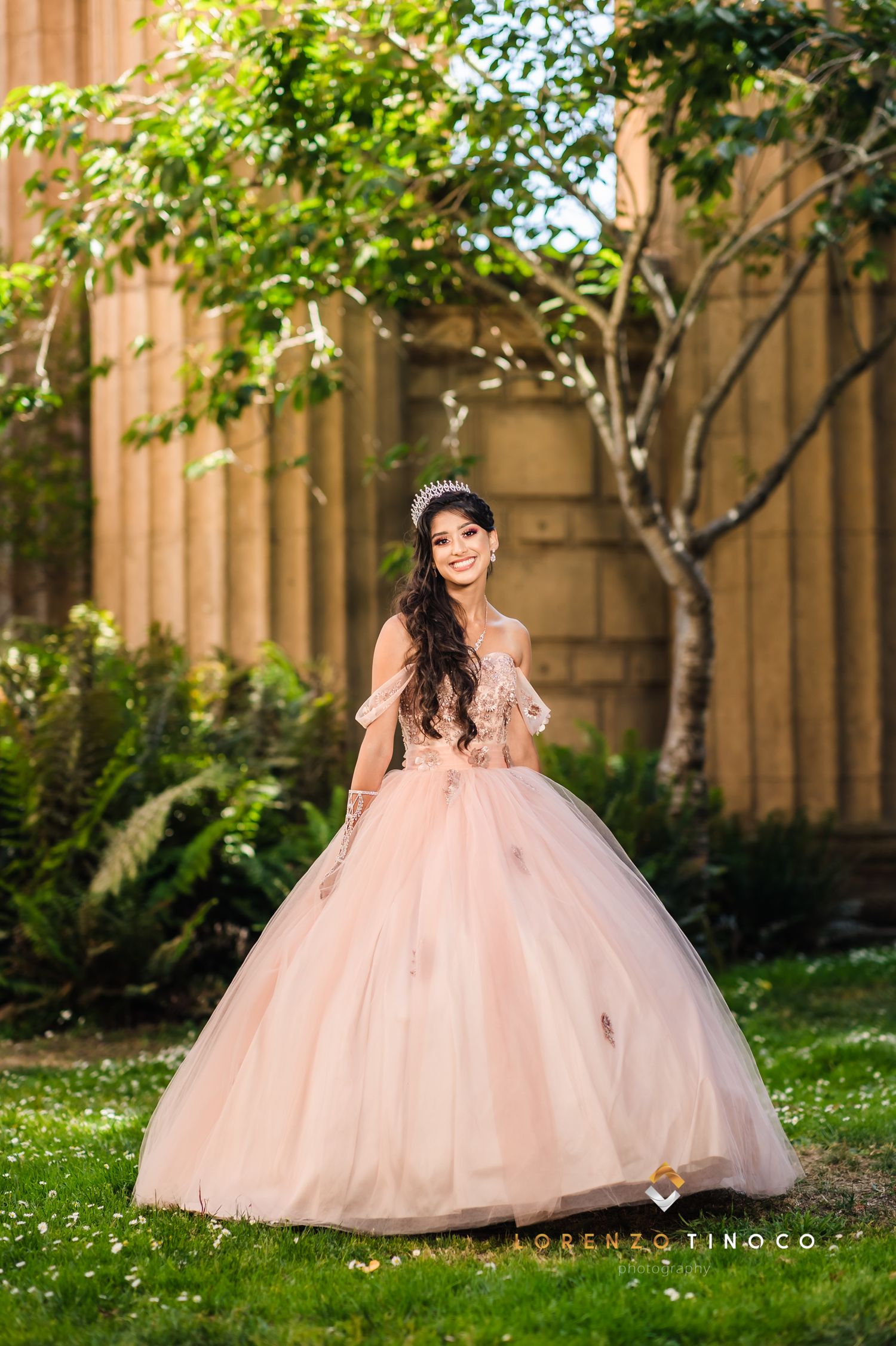 Woman in elegant pink princess gown poses against classical architecture and greenery.