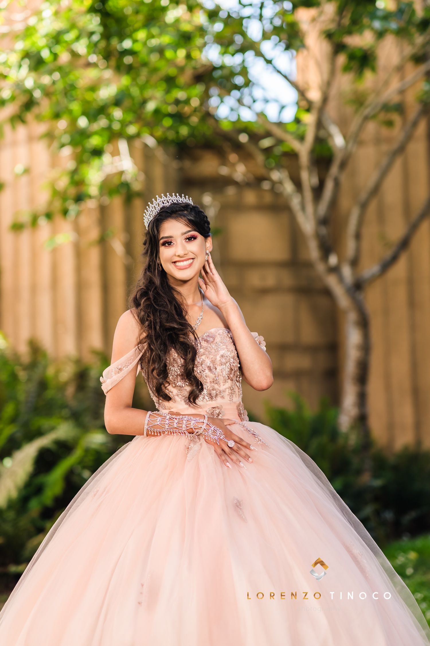 Young woman in pink tulle ballgown and tiara smiles while touching her face in an outdoor garden setting.