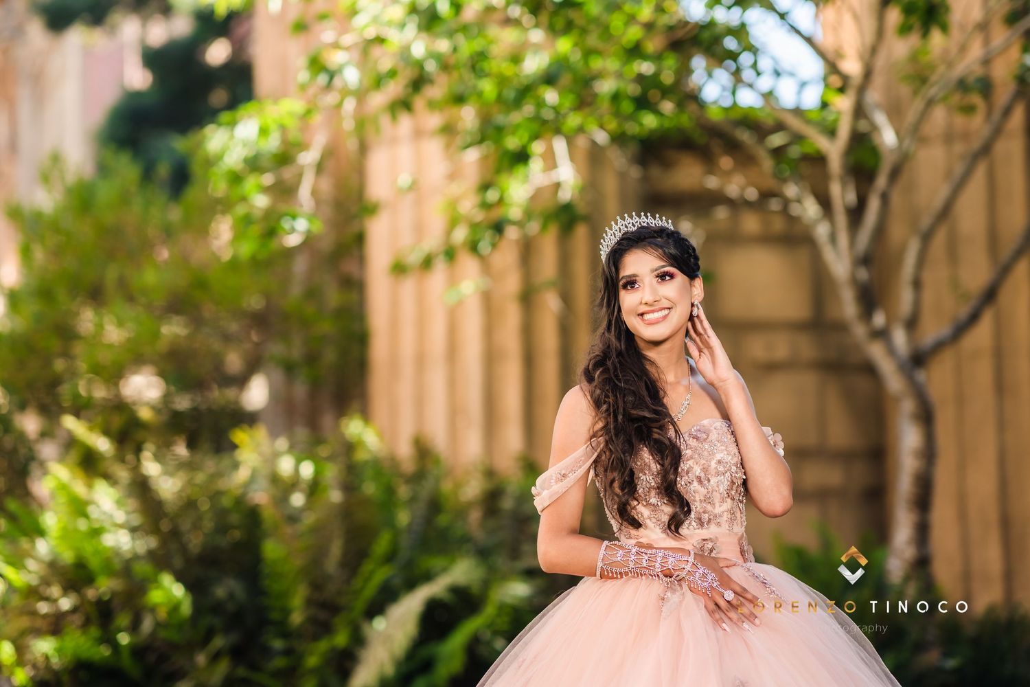 Elegant portrait of a quinceañera in a blush gown against garden foliage.