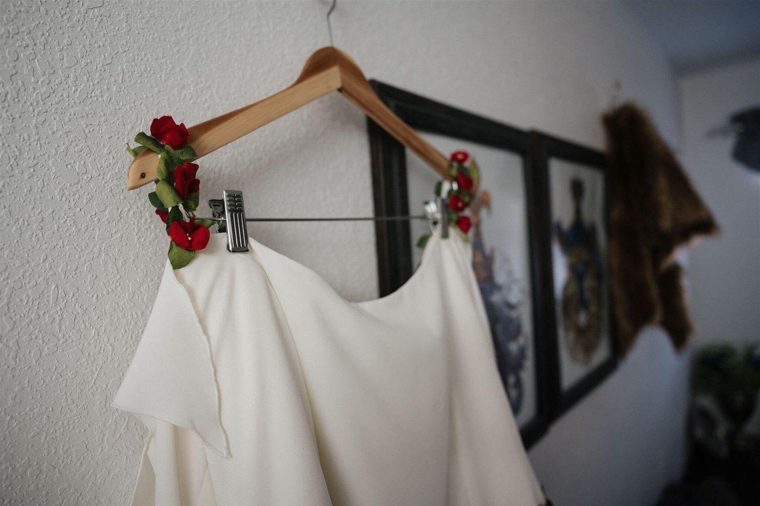 White wedding dress hanging with red flower crown detail against a white wall.