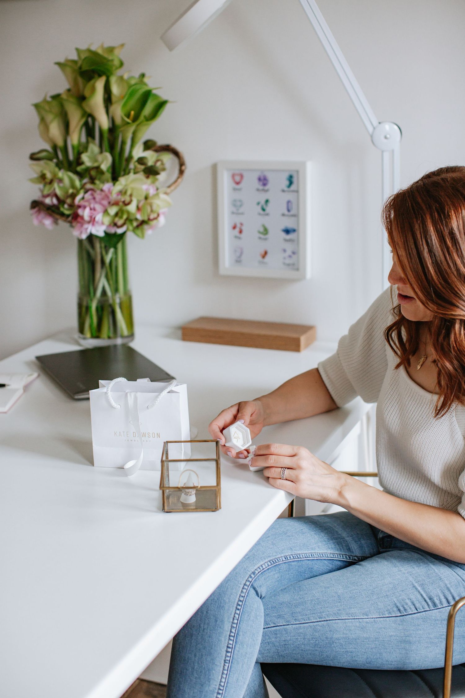Woman in casual attire working at white desk with floral arrangement and framed art.
