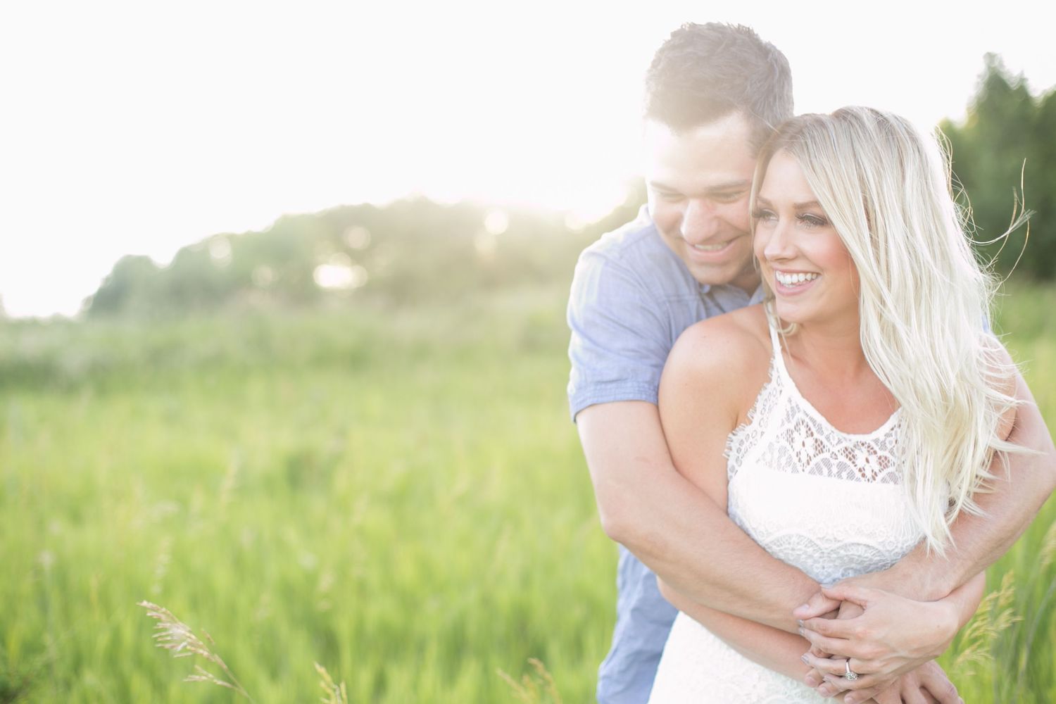 Man embracing woman from behind in glowing sunset field of green grass.