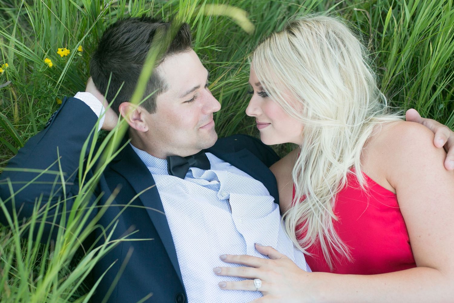 Couple lying in green grass gazing at each other during engagement shoot.