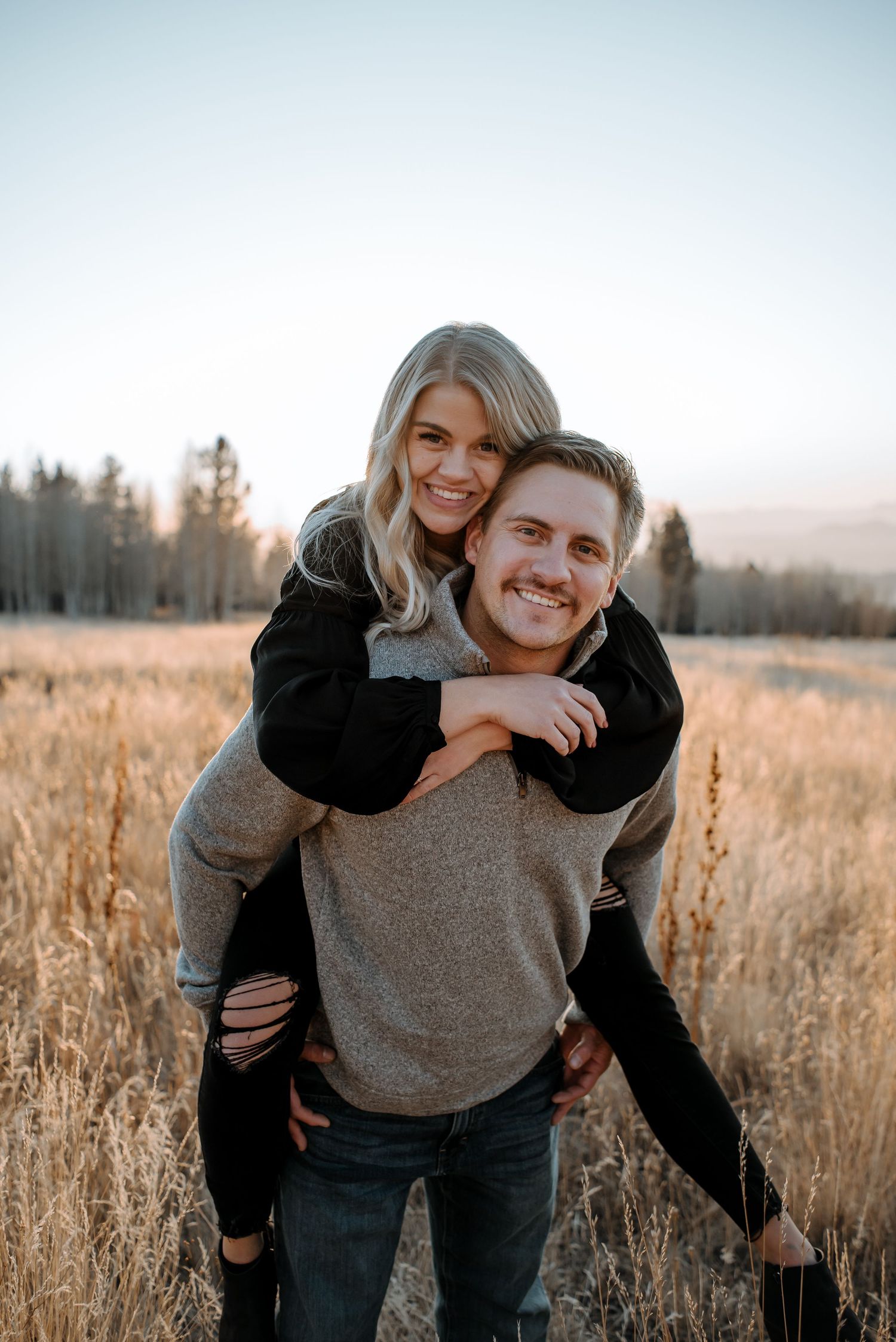 Couple enjoying piggyback ride in rustic field with sunset lighting.