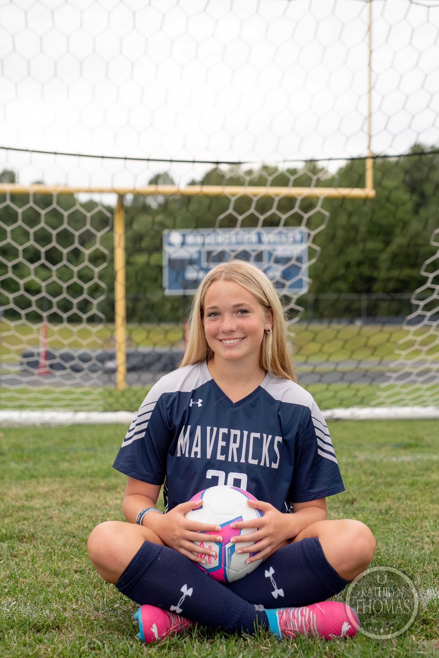 Player in Mavericks uniform sitting cross-legged with soccer ball on field.