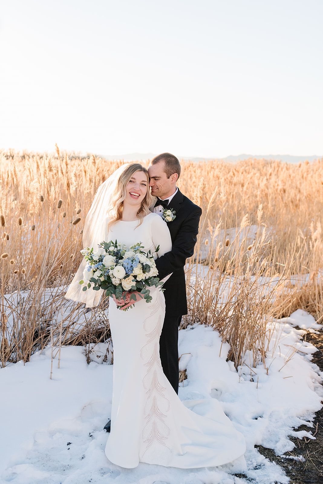 Wedding couple poses in a snowy field with golden grasses and a white floral bouquet.