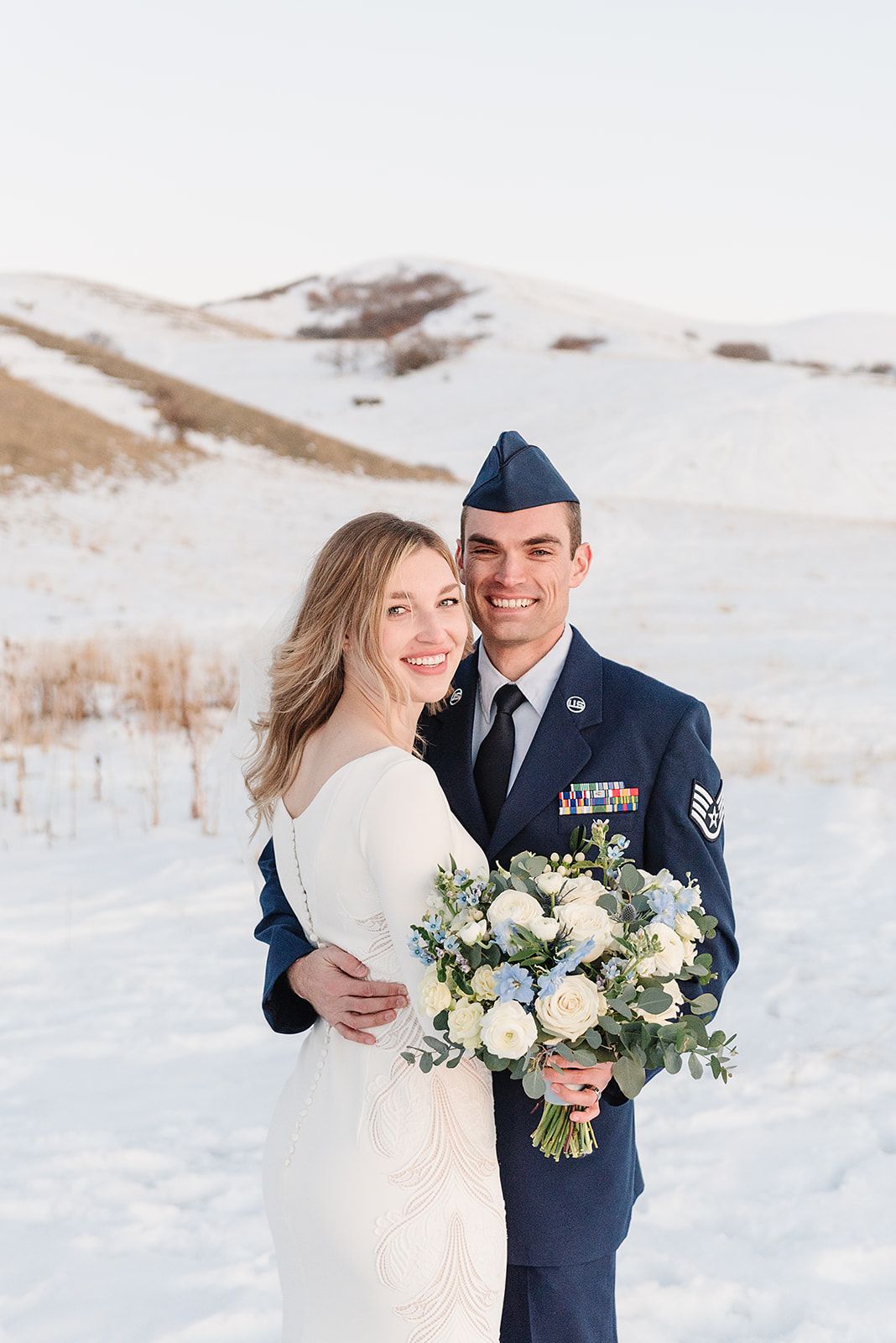 A winter wedding photo shows a couple embracing in snowy mountains with a white and blue floral bouquet.
