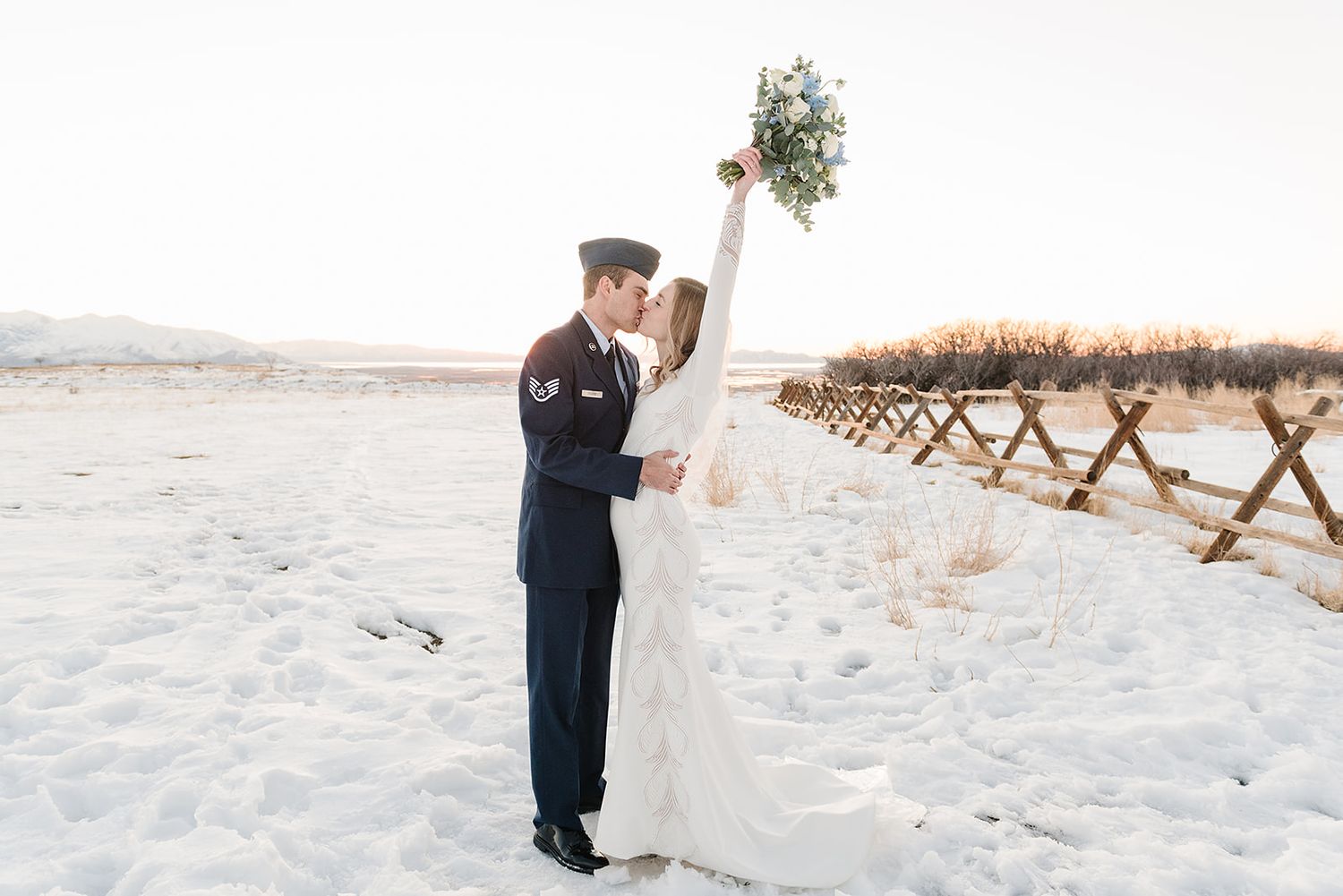 Newlyweds celebrate in the snow with a raised bouquet against a winter landscape.