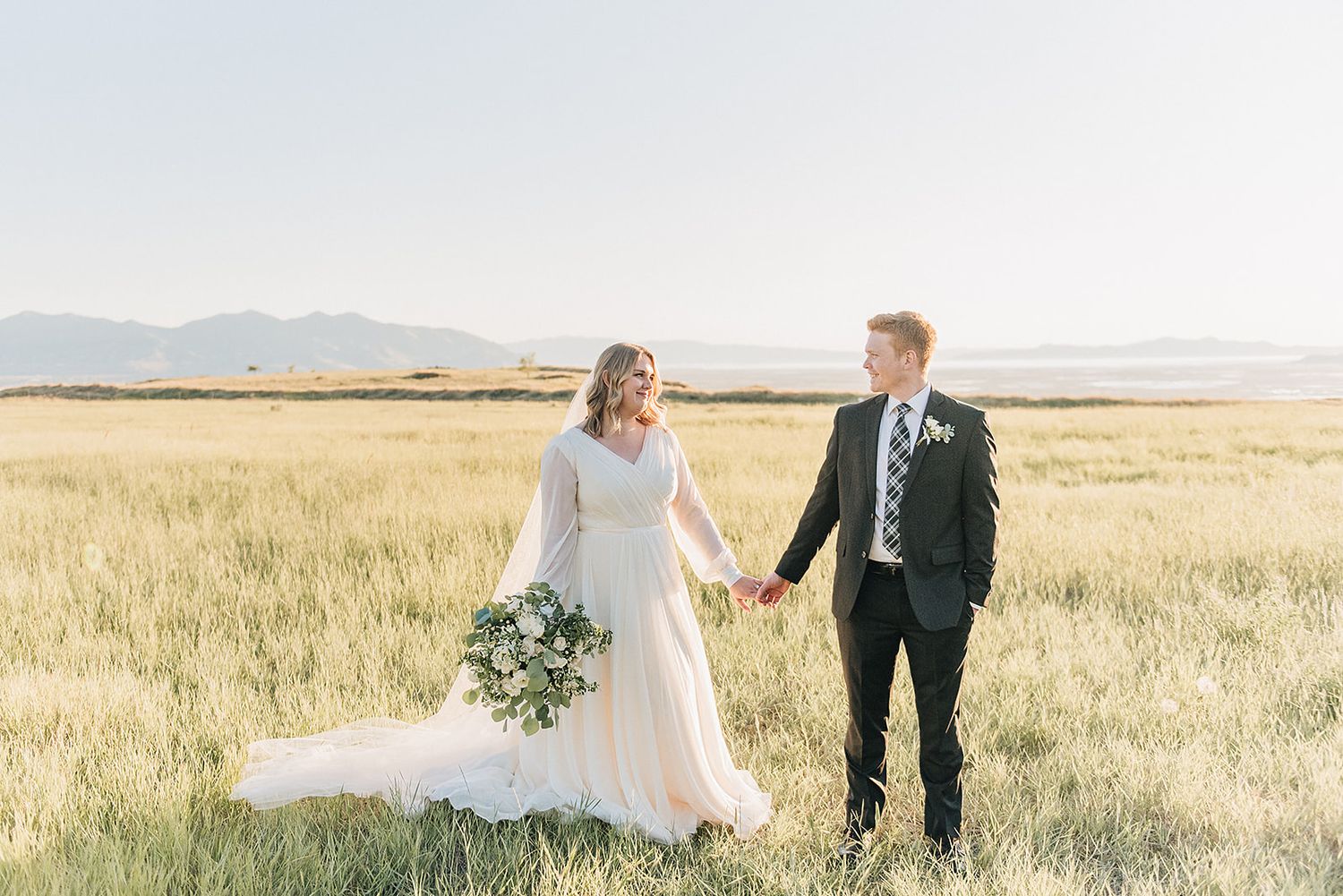 A couple stands together in a golden wheat field with mountains in the background.