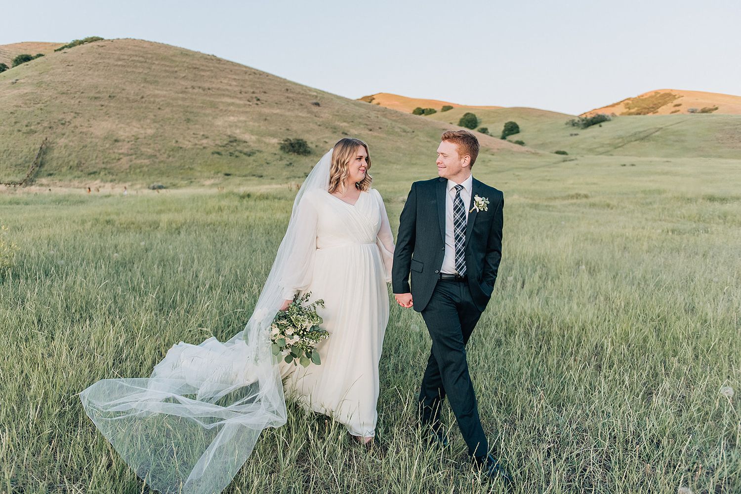 A wedding couple poses in a rolling green meadow with hills in the background.