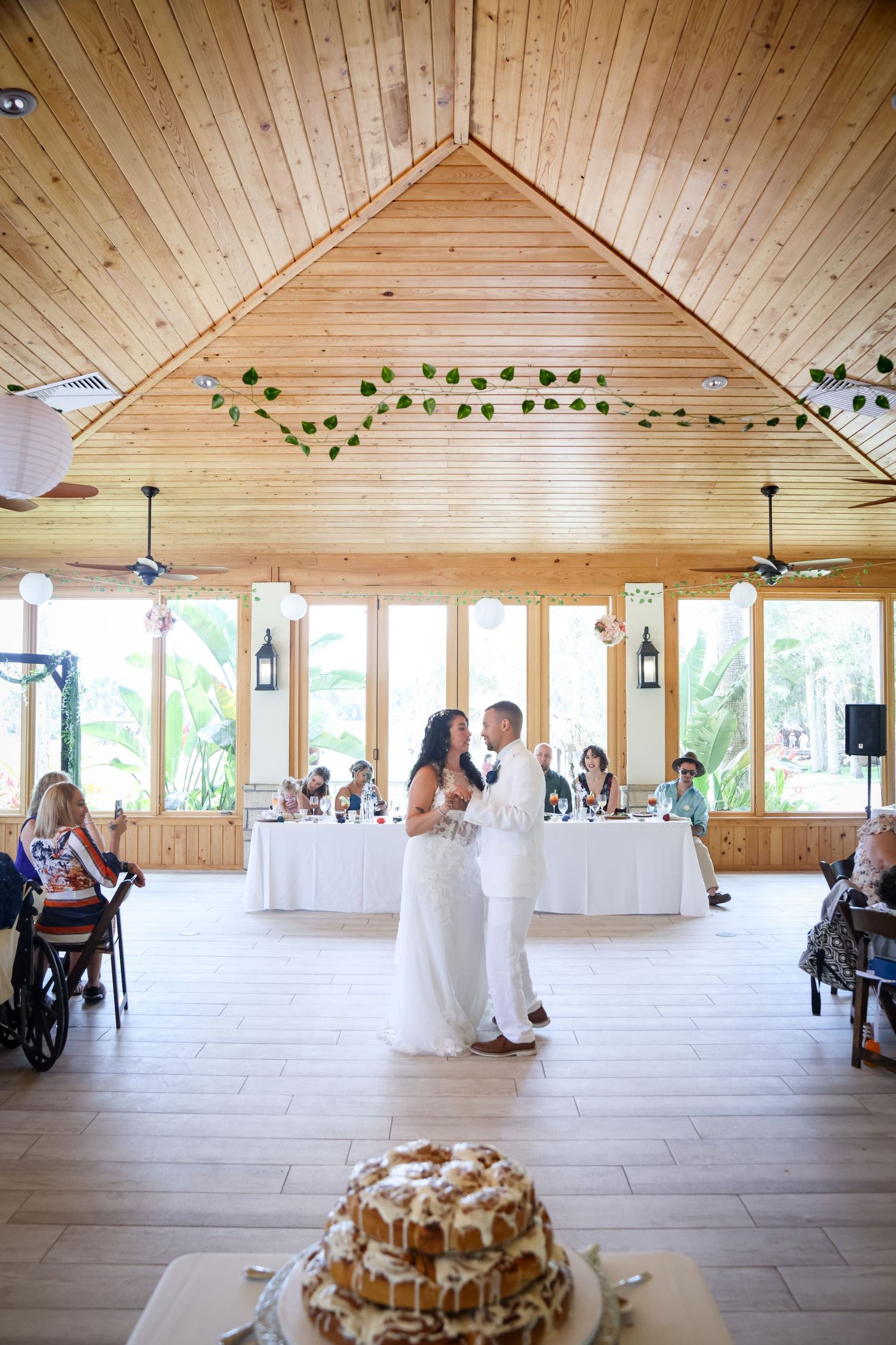 First dance in rustic wooden reception venue with decorated ceiling and wedding cake in foreground.