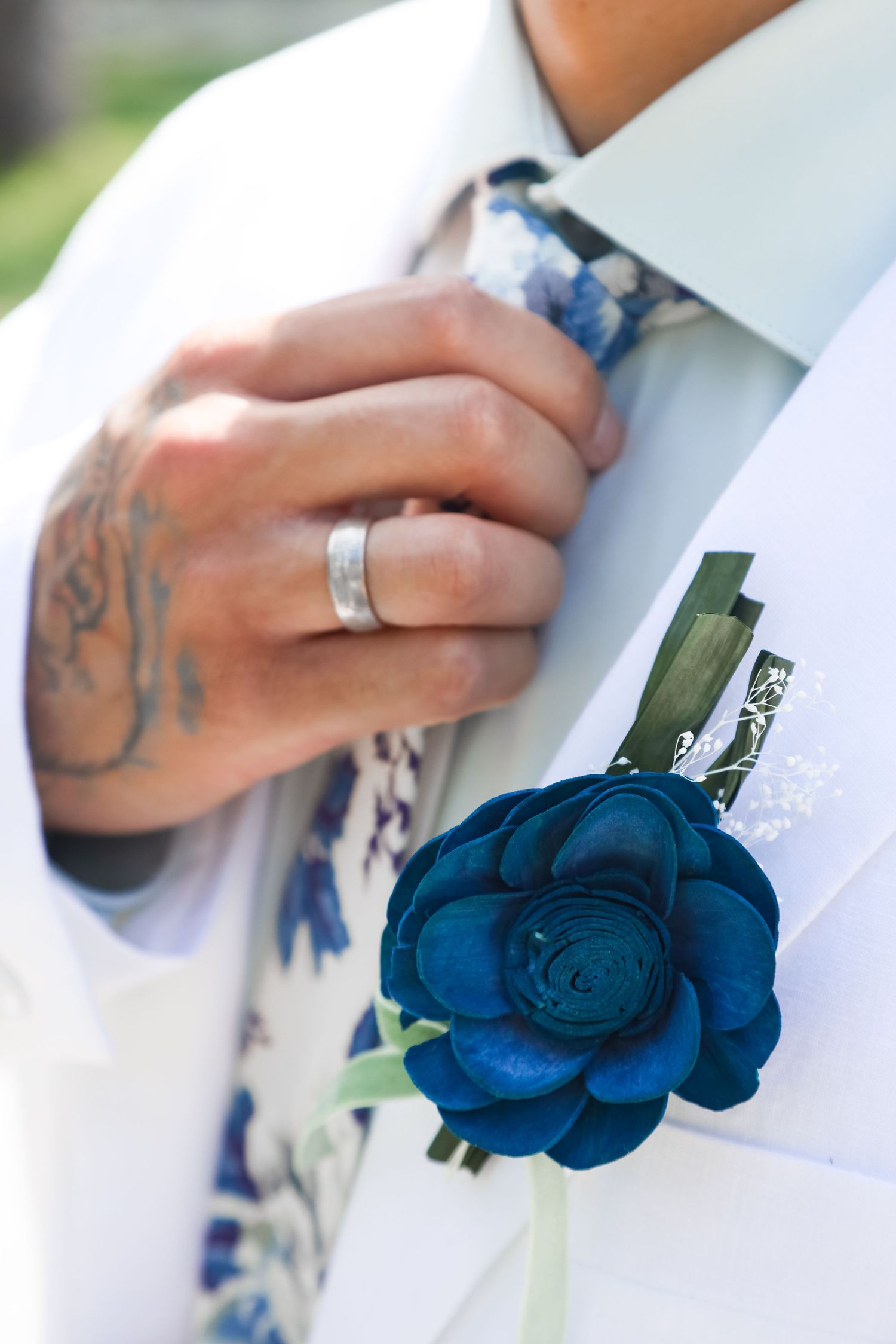 Close-up of groom's hand with ring and blue floral boutonniere on white suit.