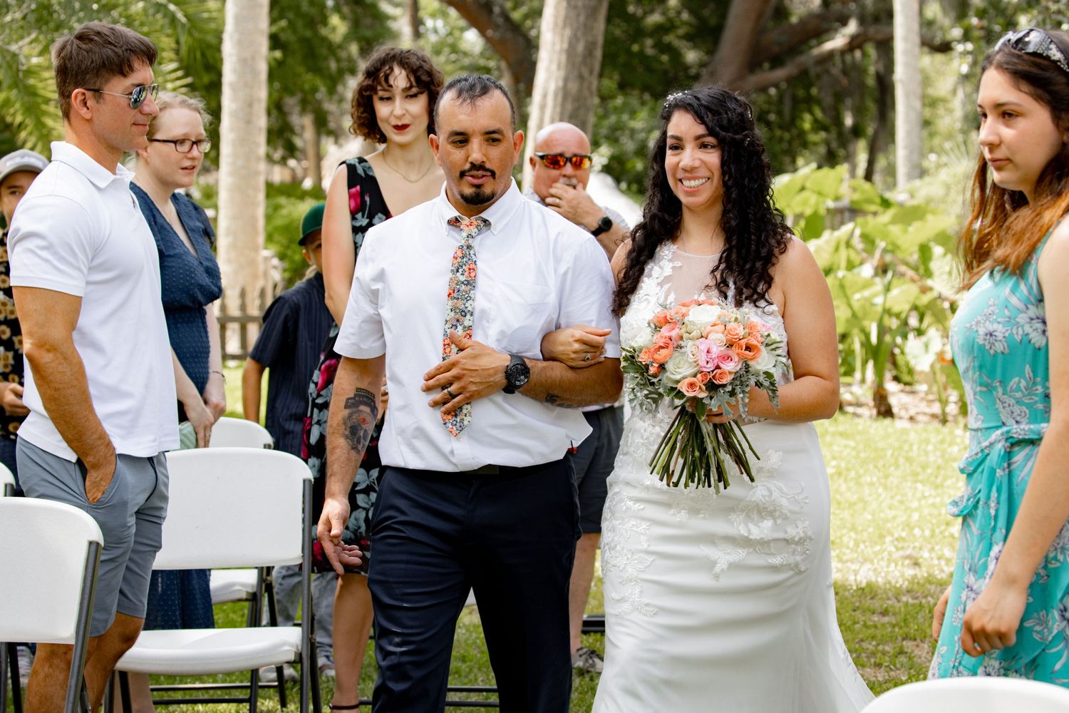 Wedding ceremony in progress under tree canopy with white chairs and floral decorations.