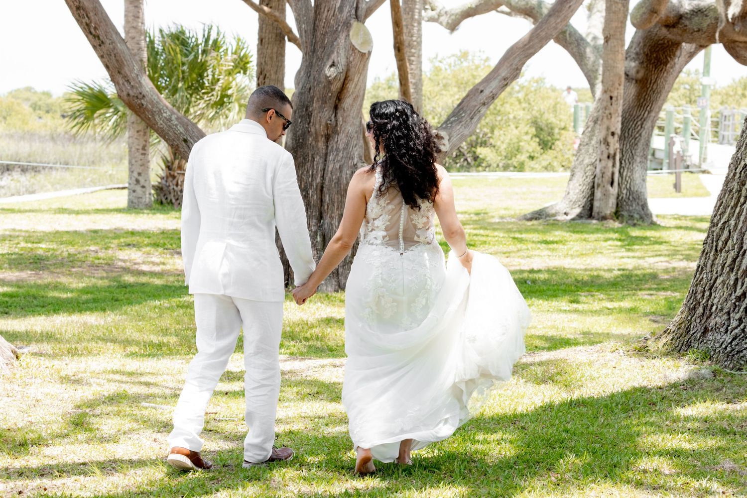 A couple in all white attire walking hand in hand through a sun-drenched garden with majestic trees.