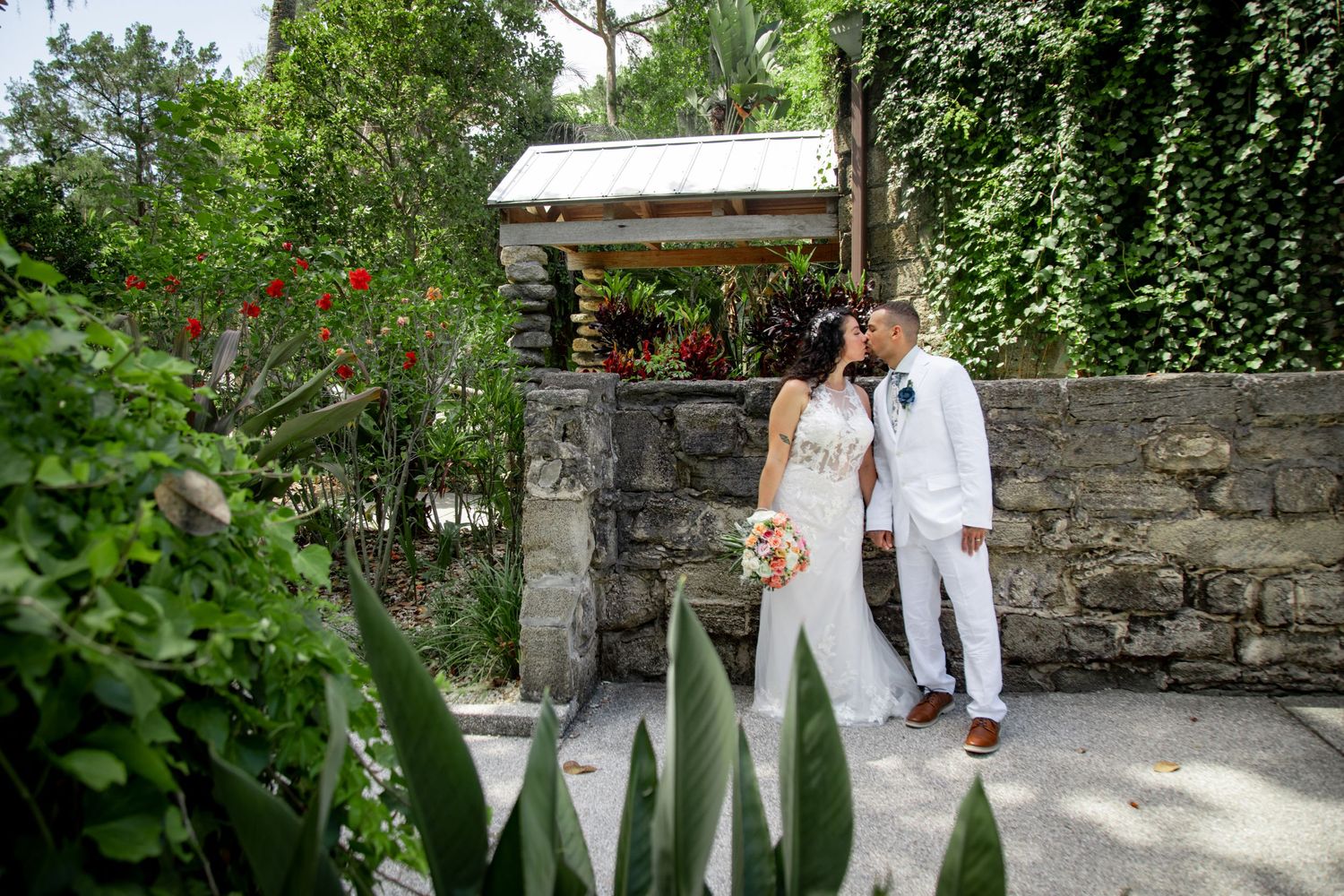 Newlyweds sharing a romantic moment by a stone wall covered in lush vines and flowers in a garden setting.