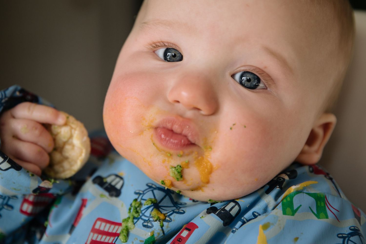 Messy baby wearing patterned bib with food around mouth enjoys eating.