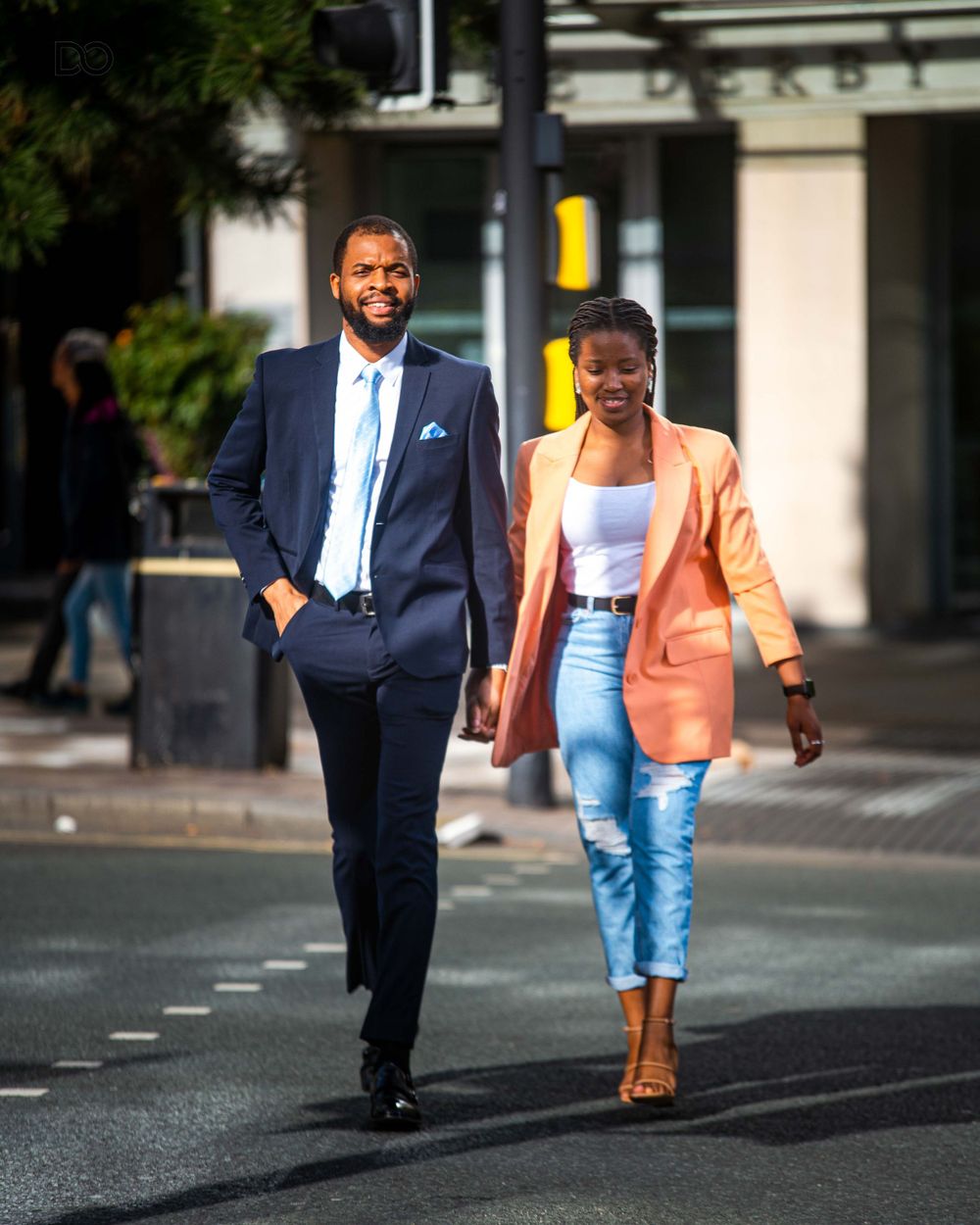 A stylish couple walks down an urban street, wearing business attire with the woman in a peach blazer and man in navy suit.