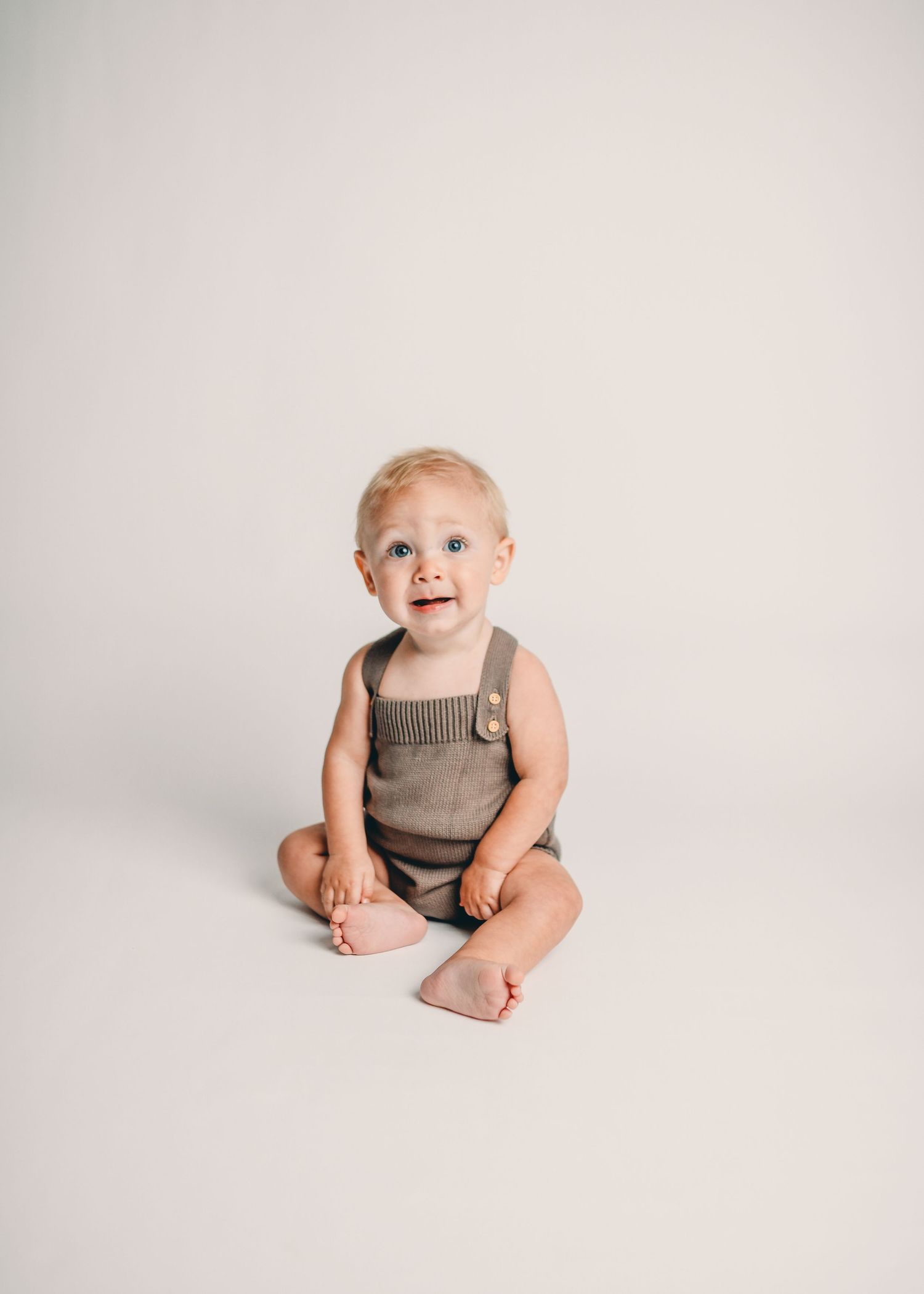Baby in tan overalls sits on white background during portrait session.