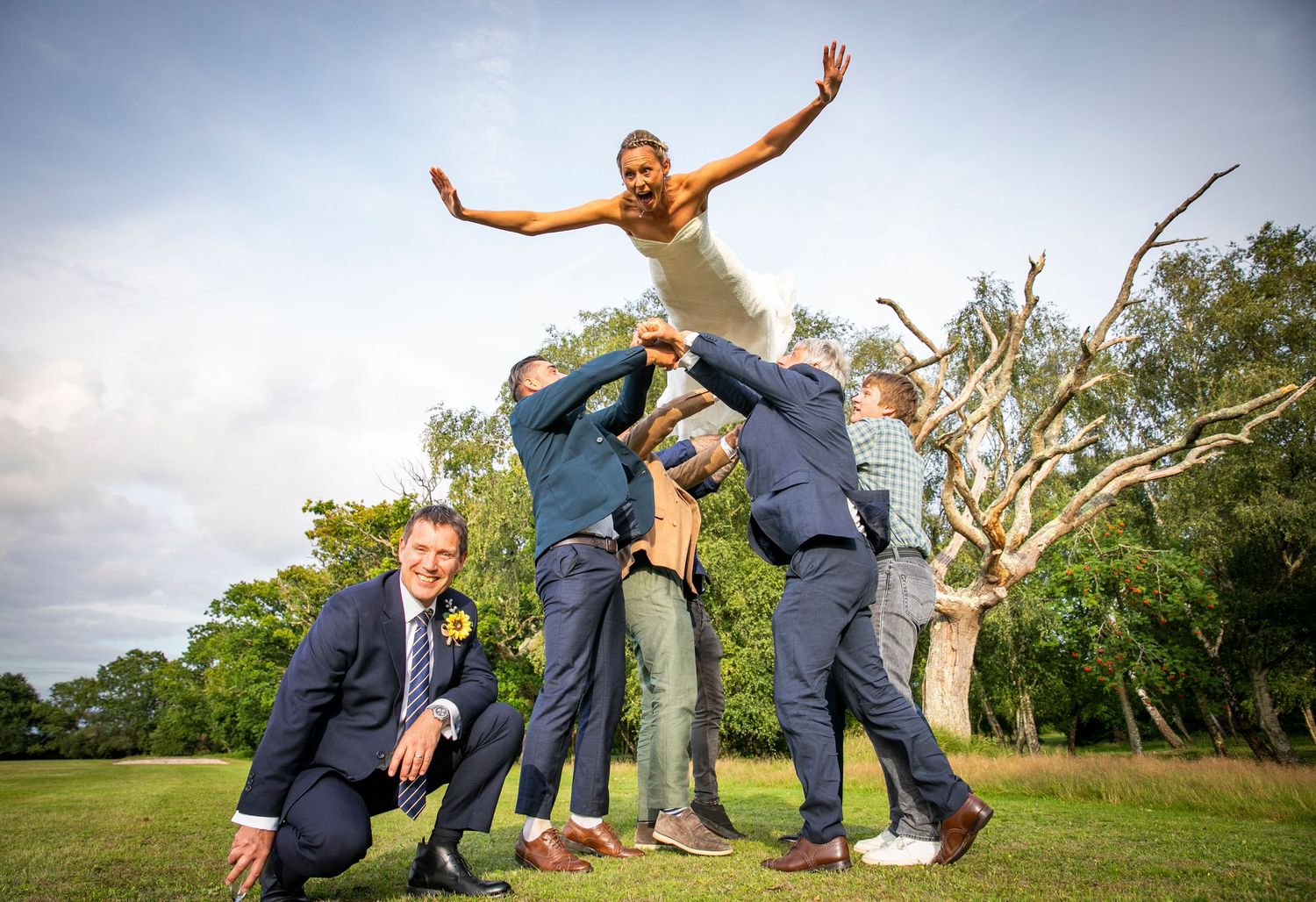 Wedding party members strike playful jumping poses in a sunny outdoor field setting.