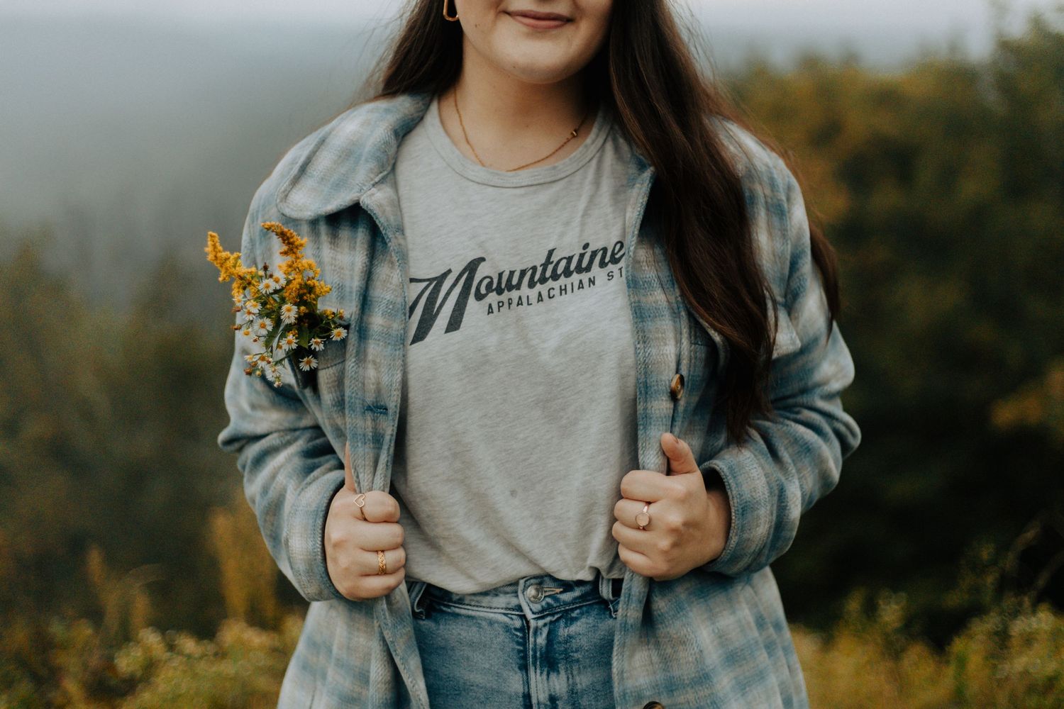 Person wearing denim jacket and Mountains t-shirt holding wildflowers outdoors.
