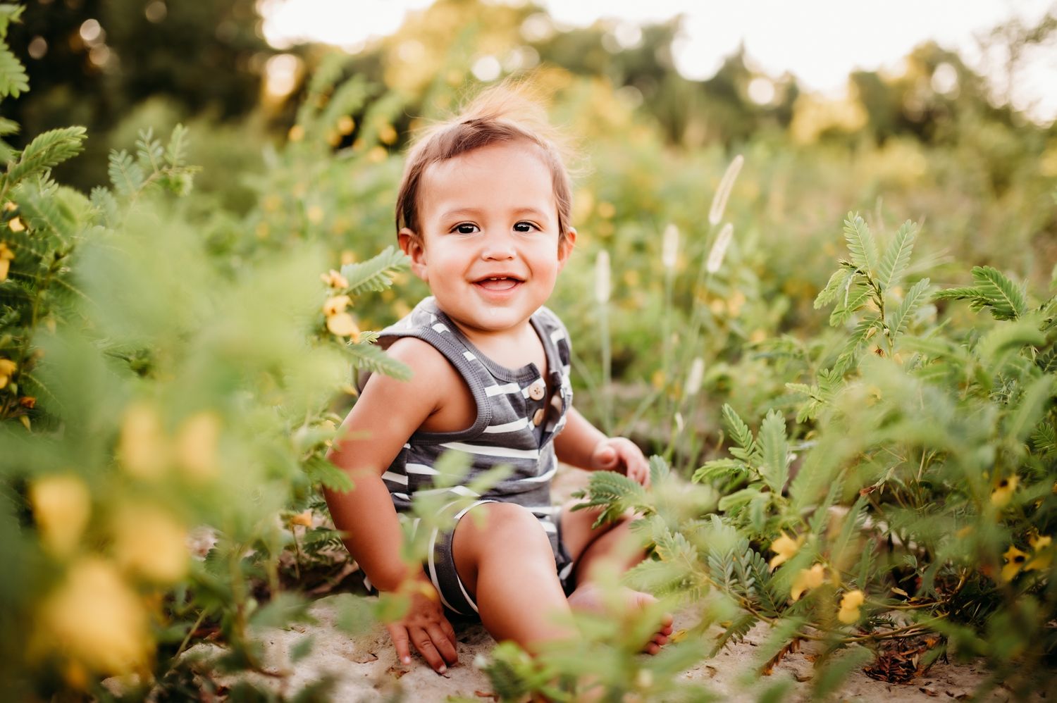 Happy toddler sitting among green plants and flowers in a summer garden.
