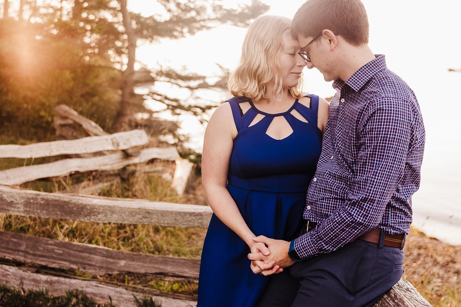 A romantic couple embraces on driftwood at sunset near the coast wearing navy blue and plaid.