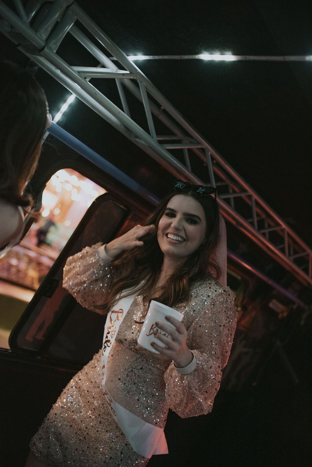 A person in a sparkly dress holds a drink while posing at a nighttime event.
