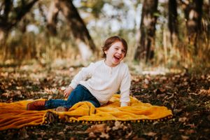 Child laughing joyfully while sitting on orange blanket in forest setting with autumn leaves.