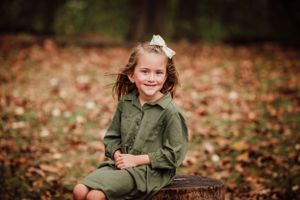 Young child wearing olive green outfit sits on wooden stump surrounded by autumn leaves on ground.