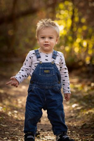 Toddler in denim overalls and printed long sleeve shirt stands on woodland path.