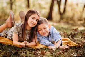 Two siblings lying on an orange blanket outdoors during autumn surrounded by fallen leaves.